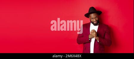 Cunning black man having an idea, smiling devious and rubbing hands, standing against red background and scheming Stock Photo