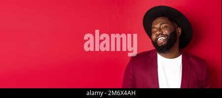 Close-up of handsome Black man with long beard laughing and having fun, looking carefree, standing over red background Stock Photo
