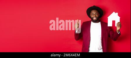 Real estate. Stylish african american bearded man showing home maket, thumb up, recommending agency or apartment complex, standi Stock Photo