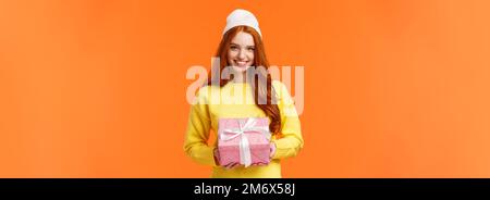 Cute redhead european woman prepare gift, wrapped present and giving it on christmas holidays, wearing winter beanie, smiling jo Stock Photo