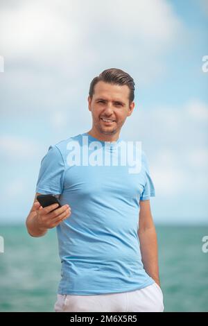 Young man holding mobile phone in his hand while walking on the beach Stock Photo