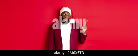 Christmas, party and holidays concept. Impressed Black man with beard, wearing santa hat, pointing finger left and gasping amaze Stock Photo