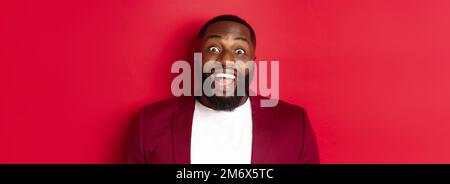 Close-up of african american man with beard staring amazed at camera, smiling and looking excited, standing in blazer against re Stock Photo