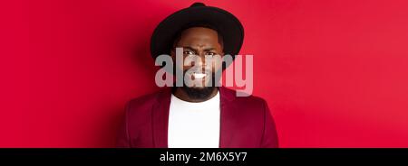 Close-up of Black man feeling uncomfortable, grimacing as watching something painful, cringe and frowning, standing over red bac Stock Photo