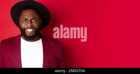 Close-up of Black man feeling uncomfortable, grimacing as watching something painful, cringe and frowning, standing over red bac Stock Photo