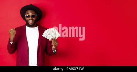 Cheerful african american guy in sunglasses, black hat and blazer, winning prize money, holding dollars and looking satisfied, s Stock Photo