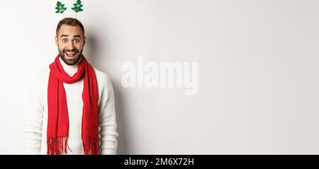 Christmas party and celebration concept. Happy bearded man looking surprised, wearing funny accessory hat, standing against whit Stock Photo