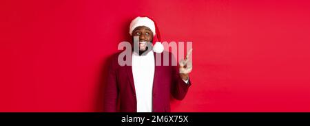 Christmas, party and holidays concept. Cheerful Black guy in santa hat pointing fingers left, showing banner, standing amused ag Stock Photo