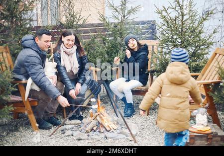 Family in warm clothes sitting around burning camp fire outside, roasting marshmallows on sticks. Stock Photo