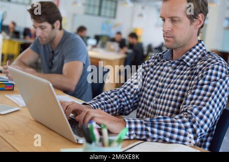 He talks code fluently. colleagues working on laptops at a desk in a large office. Stock Photo