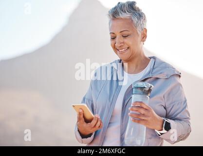 Running, water and phone with senior woman in mountain for jogging, workout and cardio training. Social media, progress tracker and fitness app with Stock Photo
