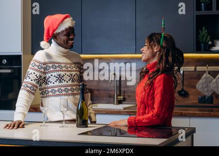 Curly-haired woman and African American man wearing festive printed sweaters drink champagne and celebrate Christmas standing in kitchen Stock Photo