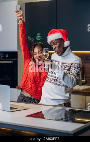 Happy young couple in festive sweaters and funny hats talks via video call using laptop and celebrates Christmas standing in kitchen Stock Photo