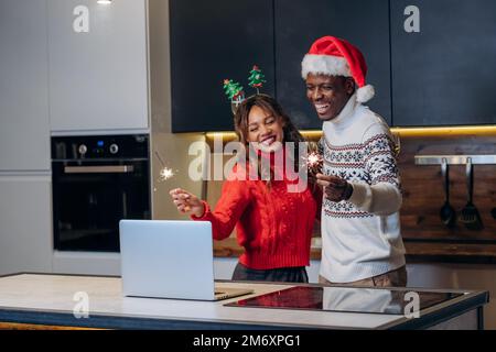 Young couple of black man and white woman communicates via video call using laptop and celebrates Christmas holding sparklers in kitchen Stock Photo