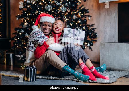 Happy woman and black man in festive sweaters hug holding gifts and sitting on carpet near Christmas tree with ornaments and garland Stock Photo