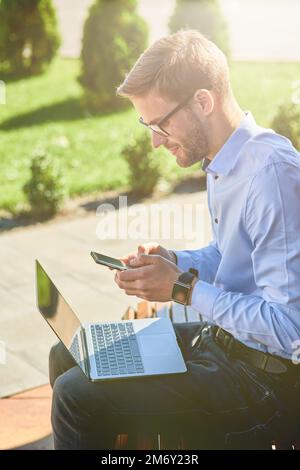 Side view of a young businessman using smartphone and working on laptop while sitting on the bench outdoors on a sunny day Stock Photo