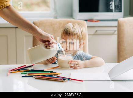A little girl has breakfast in the kitchen and draws, her mother pours milk into a mug. Stock Photo