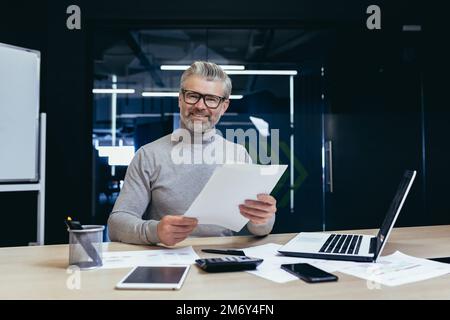 Successful businessman with documents smiling and looking at camera, portrait of mature gray-haired investor financier inside modern office, man behind paper work with reports accounts and contracts Stock Photo