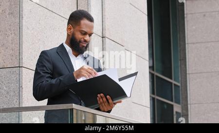 Serious African American ethnic businessman entrepreneur man employer company CEO standing on balcony in office holding folder with papers reading Stock Photo