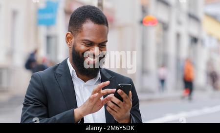 Smiling ethnic African American happy bearded man businessman executive employer with smartphone in city chatting texting has business chat reading Stock Photo