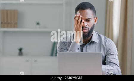 Stressed sick African American ethnic bearded man suffer with headache pain office worker businessman with laptop computer sad upset frustrated with Stock Photo