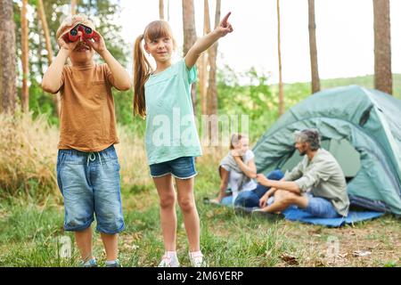 Boy looking through binoculars standing by sister pointing while camping in forest during vacation Stock Photo
