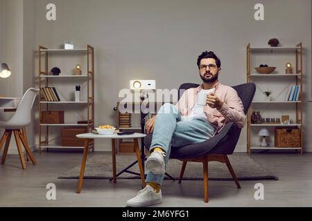 Man sitting in an armchair at home, using a projector, watching a movie, and drinking tea Stock Photo