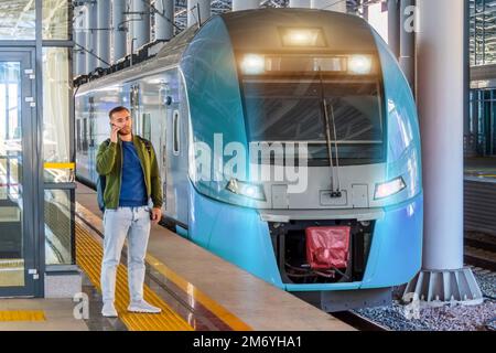 Caucasian man traveler in green windbreaker and with backpack stands on rail road platform talking on the phone, electric commuter train motion speed Stock Photo