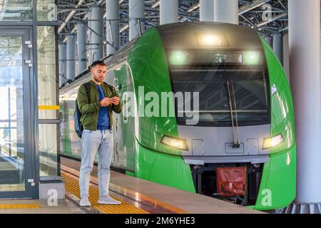 Caucasian man traveler in green windbreaker and with backpack stands on rail road platform typing text on phone, electric commuter train morning at st Stock Photo