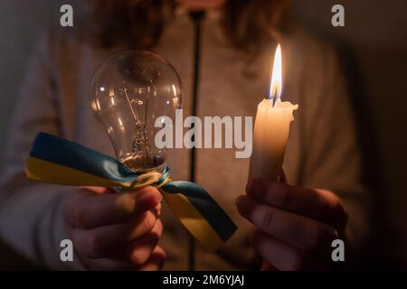 https://l450v.alamy.com/450v/2m6yjaj/blackout-power-outage-concept-girl-holds-a-burning-candle-and-an-electric-light-bulb-with-a-yellow-blue-ribbon-national-symbol-of-ukraine-2m6yjaj.jpg