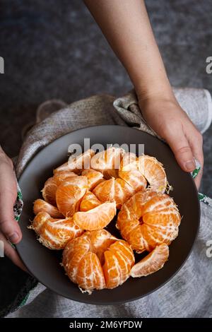 Tangerines in a bowl. The hands of a European woman hold a plate of peeled tangerines. Selective focus, dark and moody photo, shallow depth of field Stock Photo