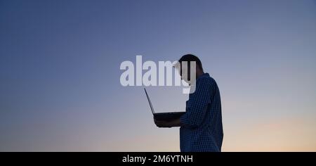 Hardly working evening: male freelancer developer or it support manager in silhouette working outdoor using laptop at sunset. Adorable dusk at background. High quality image Stock Photo