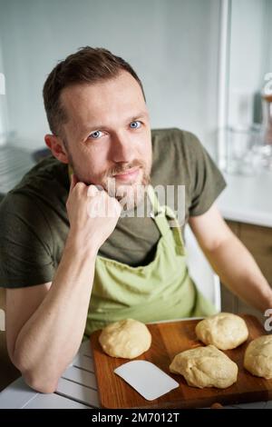 Handsome caucasian baker at home sitting at the table with dough. Portrait of man baker in green apron uniform smiling looking at camera. High quality photo Stock Photo