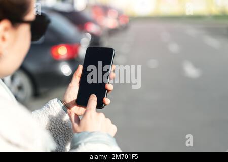 Woman using her smartphone touching screen with her finger while standing on car parking. Stock Photo