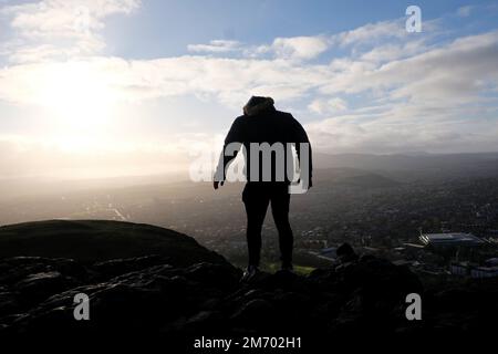 Edinburgh, Scotland, UK. 6th January 2023. Cold Blustery wind and changeable weather with some sunshine on Arthur's Seat. Leaning into the wind On top of Arthurs Seat and Silhouetted in the low winter sun, looking south towards the Pentlands.  Credit: Craig Brown/Alamy Live News Stock Photo