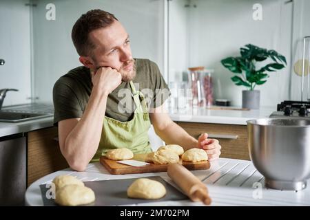 Male chef baker in green apron thinking about homemade pastry. Dough balls proofing on cutting board. Working at home kitchen concept, homemade baking. High quality image Stock Photo