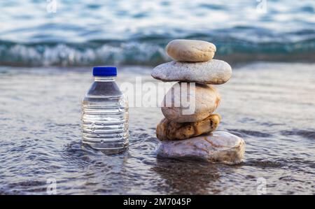 pebbles in pyramid one over another and bottle with drinking water on big rock beach sea waves in background.pure water.bottle and stones surrounded b Stock Photo