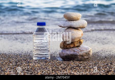 pebbles in pyramid one over another and bottle with drinking water on big rock beach sea waves in background.pure water.bottle and stones surrounded b Stock Photo