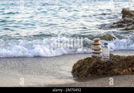 pebbles in pyramid one over another and bottle with drinking water on big rock beach sea waves in background.pure water.bottle and stones surrounded b Stock Photo