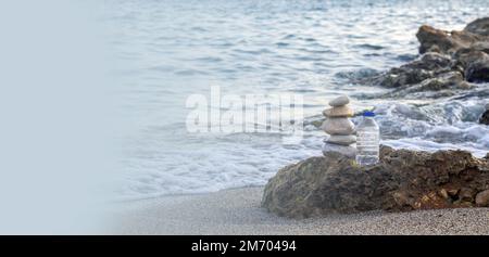 pebbles in pyramid one over another and bottle with drinking water on big rock beach sea waves in background.pure water.bottle and stones surrounded b Stock Photo