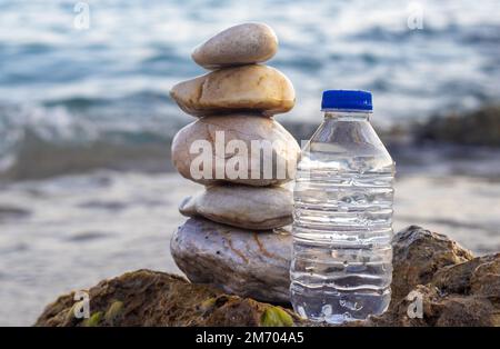 pebbles in pyramid one over another and bottle with drinking water on big rock beach sea waves in background.pure water.bottle and stones surrounded b Stock Photo