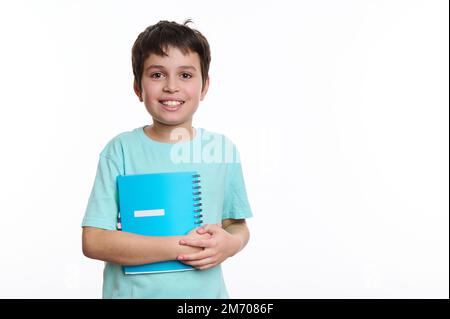 Adorable teen schoolboy in blue shirt holding a copybook and smiling a cheerful toothy smile at camera. Back to school Stock Photo