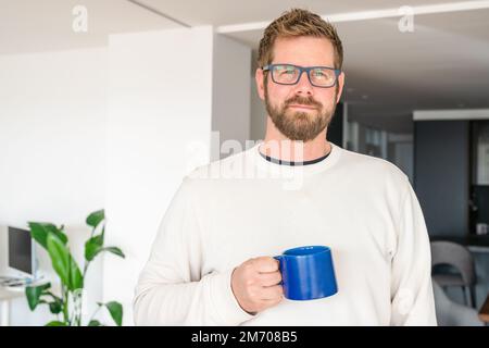 Close up of a smiling young bearded man drinking tea from a mug at the living room Stock Photo