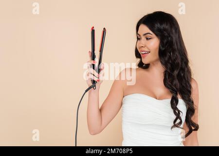 Smiling brunette woman with hairstyle looking at curling iron isolated on beige,stock image Stock Photo