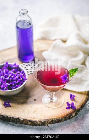 Pink cocktail in vintage glass with flower garnish, flowers and violet syrup on raw wood Stock Photo