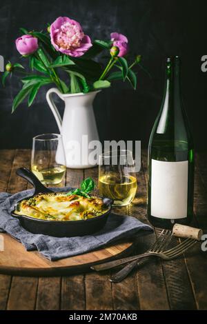 Table set with baked pasta in small cast iron skillet with wine glasses and bottle and flowers Stock Photo