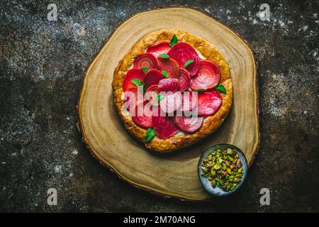 Round pastry tart with sliced beets, feta and mint leaves on wooden board with pistachios in bowl Stock Photo