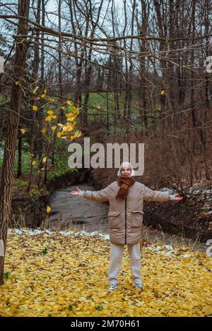 the girl rejoices in autumn and throws yellow leaves; wearing a jacket, a hood from a hoodie and a scarf, wearing sneakers Stock Photo