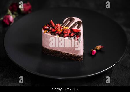 Heart cake decorated with pomegranate and strawberries. Dessert on a black plate with dried rose. Stock Photo