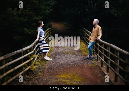 A romantic photo of a young couple in a beautiful autumn park, standing on opposite sides of a pathway and leaning on the railing Stock Photo